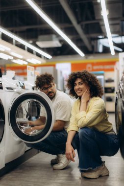 Bir erkek ve kadın, modern bir elektronik mağazasında çamaşır makinesiyle etkileşime geçip seçenekleri araştırıyorlar..