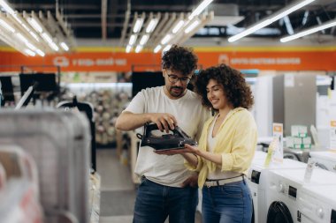 Elektronik mağazasında genç bir çift birlikte yeni bir ütüye bakıyor, alışveriş yaparken ilgi ve takım çalışması sergiliyorlar..