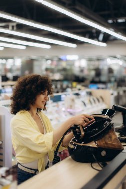 Sarı bluzlu bir kadın, teknolojik olarak gelişmiş bir elektronik mağazasında çeşitli aletlerin arasında buhar ütüsünü inceliyor..