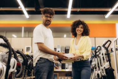 İki kişi bir elektronik mağazasını keşfediyor, çeşitli seçenekler arasından gülümseyerek bir cihaz seçiyorlar..