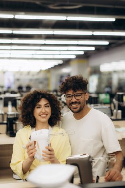 Modern bir elektronik mağazasında mutfak aletlerini keşfeden neşeli bir çift, birlikte alışveriş yaparken mutluluk ve heyecan ifade ediyorlar..