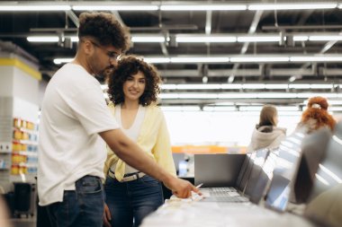 İki birey, teknoloji ve karar verme mekanizmasını simgeleyen parlak ışıklı bir elektronik mağazasında dizüstü bilgisayarları inceliyor..