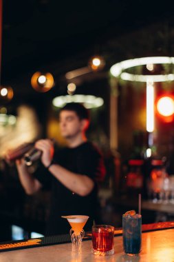 Bir barmen, modern bir barda renkli bir aydınlatmayla kokteyl sallarken görülüyor. Yanında da tezgahta üç kokteyl var..