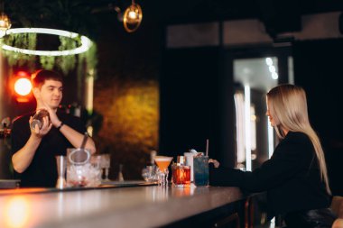 Bir barmen barın arkasında içkileri ustalıkla hazırlarken bir müşteri de akşamdan zevk alır. Ortam aydınlatma ve sofistike dekor, sosyalleşme ve rahatlama için sıcak, canlı bir atmosfer yaratır..