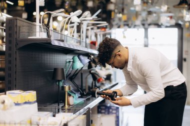 Beyaz gömlekli bir adam aydınlatma mağazasında çeşitli aydınlatma donanımı ve aksesuarlarıyla çevrili bir masa lambasını inceliyor. Dekor, ev veya ofis dekorasyonu için çağdaş perakende ve alışveriş çıkarlarını yansıtıyor.