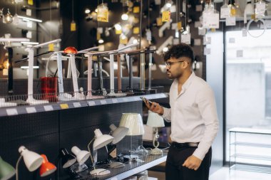 Bir adam, çeşitli lamba tasarımları, renkler ve sergilenen stillerle çevrili telefonuna bakarken bir aydınlatma mağazasındaki lamba seçimine göz atar..