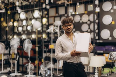 Gözlüklü ve beyaz gömlekli bir adam, modern bir aydınlatma mağazasında çeşitli aydınlatma aletleri sergilerken elinde bir kutu tutuyor..