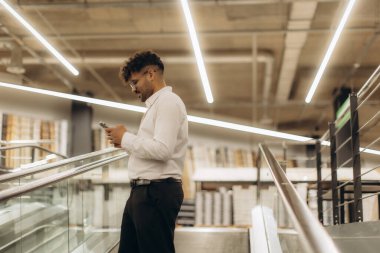 Çağdaş bir çalışma alanındaki yürüyen merdivende dururken akıllı telefonunu kontrol eden beyaz tişörtlü genç adam profesyonel bir ortamda iletişimi ve teknolojiyi gösteriyor..