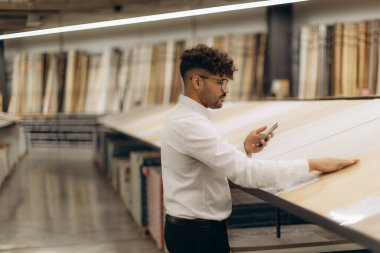 Beyaz gömlekli bir adam ahşap panelleri inceliyor ve ev geliştirme malzemeleri ve döşeme ürünleri konusunda uzmanlaşmış geniş, iyi ışıklandırılmış bir mağazada seçenekleri karşılaştırmak için telefonunu kullanıyor..