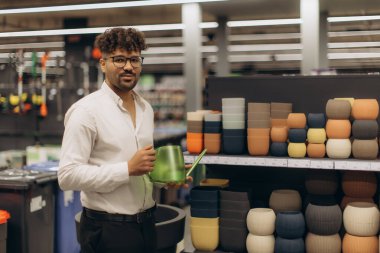 Bir mağazanın bahçe reyonunda elinde yeşil bir su kovası ve arka planda renkli saksılar tutan bir adam..