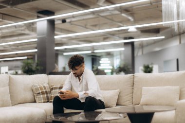 Şık bir ofis ortamında, modern iş kültürü ve rahatlamayı tasvir eden şık endüstriyel dekorlarla çevrelenmiş bir şekilde koltukta oturup mobil bir cihaz kullanan genç bir profesyonel. Tarafsız tonlar boşluğa hakim.