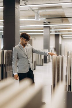 İş kıyafeti giymiş şık bir adam modern bir sergi ortamında panelleri inceliyor. Çevre, profesyonel veya kişisel projeler için çeşitli doku ve tasarım seçenekleri sunan iyi ışıklandırılmış bir ortamdır..