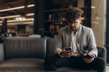 Takım elbiseli ve gözlüklü bir adam koltukta oturuyor, akıllı telefonuyla modern ve şık bir ofis ortamında meşgul, profesyonellik saçıyor ve odaklanıyor..