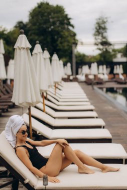 Havuz kenarında dinlenen bir turist, kafasında havluyla yaz tatilinin tadını çıkarıyor, güneşin altında ferahlatıcı bir kokteyl yudumluyor.