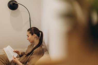 Huzurlu bir sahne, rahat bir şekilde oturan ve sıcak, aydınlık bir yaşam alanında kitap okuyan genç bir kadın..