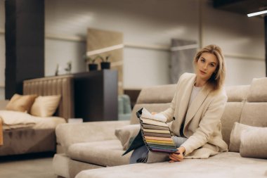 Şık bir kadın, modern bir mobilya dükkanında kumaş örneklerini değerlendirirken kanepeye oturmuş sofistike ve seçici bir görünüm sergiliyor. İç tasarım, alışveriş ve ev estetiğini temsil etmek için ideal..