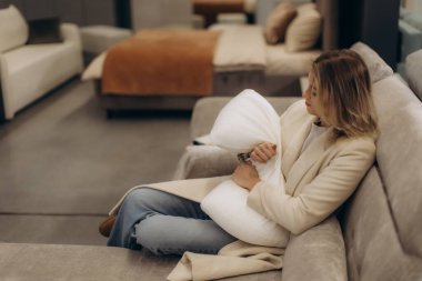 Rahat bir koltukta oturan, modern bir mobilya galerisinde yastığa sarılan bir kadın. Odak noktası rahatlama, ev konforu ve şık iç tasarım..