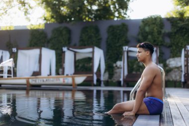 Genç erkek yüzücü antrenmandan sonra havuz başında dinleniyor, suyun sakinliğinin tadını çıkarıyor ve bir sonraki yüzüşüne hazırlanıyor.