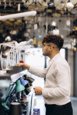 Bir adam aydınlatma ve elektrikçide masa lambaları seçerken görülüyor. Ayar, modern bir sahnede bir alışveriş etkinliğini yansıtan çeşitli ışıklandırma ürünleri sergiliyor..