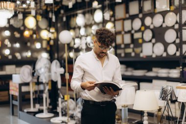 Resmi giyinmiş bir adam bir aydınlatma mağazasını keşfederken kitap okuyor. Arka plan, çeşitli lambalar, avizeler ve modern aydınlatma ürünlerinin parlak bir şekilde ışıklandırılmış görüntülerini gösteriyor..