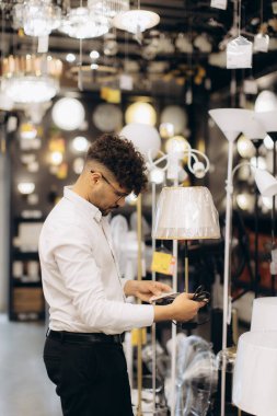 Beyaz gömlekli bir adam, şık dekorasyonlar ve aydınlatılmış ürünlerle dolu bir aydınlatma mağazasında ev ve ofis kullanımı için çeşitli modern ve geleneksel tarzlar sergileyen bir lambayı inceliyor..