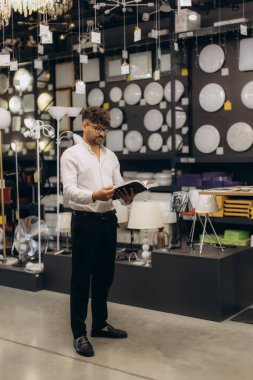 Modern bir aydınlatma mağazasında çeşitli aydınlatma armatürleri ve dekoratif lambalarla çevrili bir kataloğu inceleyen profesyonel kişi, çağdaş bir tasarım ve ev hayatı odaklı bir ortam sunuyor..