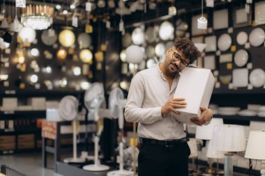Neşeli bir adam, çeşitli ışıklandırma ürünleriyle dolu iyi ışıklandırılmış bir galeride duruyor, nazikçe bir kutu tutuyor ve coşkuyla gülümsüyor..