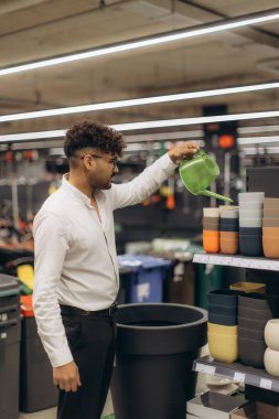 Bir adam dükkanda geziniyor, bahçe aletleri ve saksıları keşfediyor, elinde yeşil bir su kovası tutuyor. Olay yeri, düzgünce düzenlenmiş bir mağazanın içindeki bahçe malzemeleriyle eğlence, alışveriş ve ilgiyi yansıtıyor..