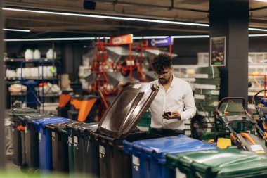 Çeşitli ürünlerin bulunduğu bir nalbur dükkanında sergilenen büyük geri dönüşüm kutularını inceleyen bir adam.