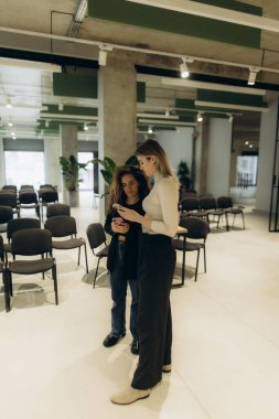 Modern bir ofiste, minimalist dekor ve bol doğal ışık içeren iki kadın sohbet ederken görülüyor. Çevre işbirliği, yaratıcılık ve profesyonel ama rahat bir atmosfer öneriyor..