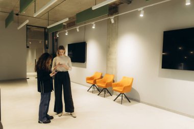 Modern ofis ortamında şık sandalyeler ve dekorlarla etkileşime giren iki meslektaş, çağdaş profesyonel ortam sergiliyor..