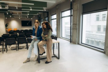 İki kadın, modern bir ofiste arka planda büyük pencereleri ve sandalyeleri olan belgeleri tutarak profesyonel bir tartışmaya giriştiler..