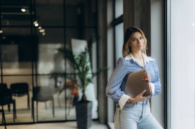 Kendine güvenen bir kadın modern bir ofisin penceresinde elinde dizüstü bilgisayarla duruyor ve gündelik ama profesyonel giysiler giyiyor. İç mekan, profesyonel ve şık bir atmosfer yayarak çağdaş dekorlar sergiliyor..