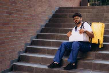 Teslimatçı adam merdivenlerde dinleniyor, elinde bir paketle oturuyor, sarı bir sırt çantası takıyor, kısa bir süre dinlenmenin tadını çıkarıyor.