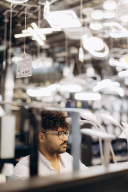 Çeşitli modern lamba tasarımları ve aydınlatmalarla çevrili bir perakende mağazasının içindeki çeşitli aydınlatma tesisatlarını keşfeden bir adam. Ayar, mevcut ışıklandırma seçeneklerinin zarif ve işlevsel çeşitlerini vurgular.