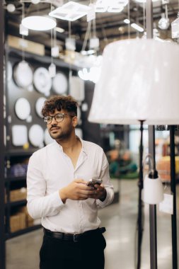 Modern bir aydınlatma mağazasında gözlüklü ve beyaz gömlekli bir adam akıllı telefon tutuyor. Çağdaş aydınlatma armatürleri ile çevrelenmiş, düşünceli ve seçici ürünlerle meşgul görünüyor..