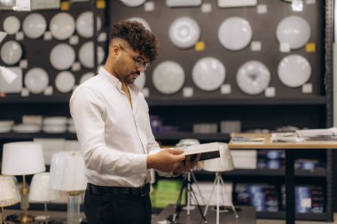 Beyaz gömlekli bir adam modern bir aydınlatma mağazasında çeşitli lambalar ve aksesuarlarla çevrili ürün detaylarını inceliyor..