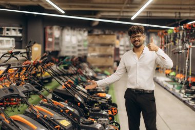 Kendine güvenen satış elemanı bir mağazada çim biçme makinesinin yanında duruyor, neşeyle gülümsüyor, mevcut modelleri sergiliyor, potansiyel müşterilere ürün özelliklerini vurguluyor, iş ortamında müşteri katılımını destekliyor..