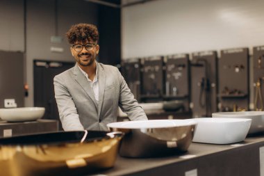 Takım elbiseli şık bir adam modern bir galeride, lüks banyo lavabolarını ve yüksek kaliteli iç dekorasyonları sergileyerek güvenle gülümsüyor..