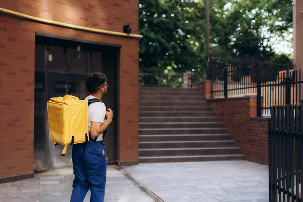 Delivery man wearing blue overalls and carrying yellow insulated backpack walking near building entrance
