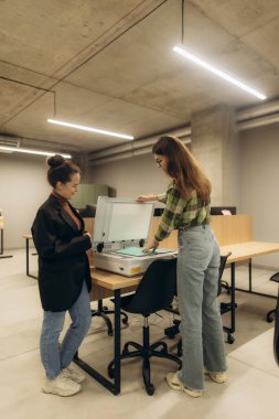 İki kadın, modern endüstriyel tarzda bir iş yerinde çalışırken bir ofis tarayıcısıyla etkileşime geçerek, işbirlikçi ve profesyonel bir çevreye işaret ediyorlar..