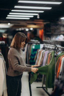 Genç bir kadın, bir moda mağazasında yeşil bir kazağı incelerken, kıyafet alışverişine özen gösterir. Parlak ışıklandırma ve canlı renkler modern ve davetkar bir perakende atmosfer sağlar.