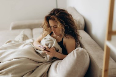 Genç bir kadın kanepenin altında oturuyor, elinde akıllı bir telefon, modern ve rahat bir oturma odasında yumuşak ışıklandırmayla rahat ve rahat bir ortamda vakit geçiriyor..