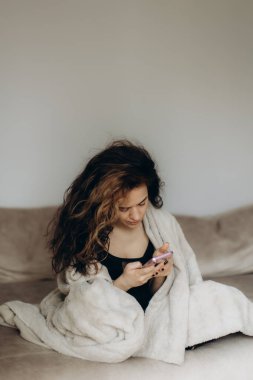 Yumuşak bir battaniyeye sarılı genç bir kadın akıllı telefonunu kullanırken kanepeye oturur. İçeride dinleniyor, odaklanmış ve rahat görünüyor, rahat ve sakin bir atmosfer yaratıyor..