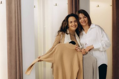 İki neşeli kadın şık bir giyim mağazasında ellerinde bej ve gri kazaklar, gülümseyerek ve neşeyle poz vererek rahatlamış bir alışveriş ortamında arkadaşlığı ve modern moda eğilimlerini temsil ediyorlar..