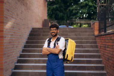Mavi tulumlu ve sarı izotermal sırt çantalı kurye çapraz kollu merdivenlerde duruyor.