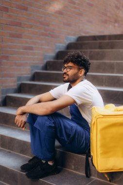 Kurye şehir merkezindeki yemek teslimatını tamamladıktan sonra sarı yalıtılmış sırt çantasıyla dışarıda dinleniyor.