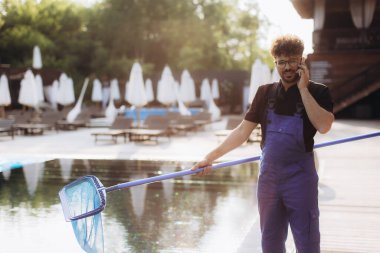 Havuz temizleyicisi mavi tulum giyip elinde yaprak tutarak lüks bir otelin havuz kenarında konuşuyor.