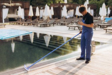 Havuz temizleyicisi mavi üniformalı otel havuzunu fırçayla temizliyor ve yaz günü akıllı telefon kullanıyor.