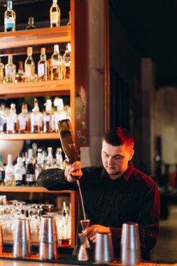Çağdaş bir bar ortamında barmen ustalıkla içki hazırlıyor. Aydınlatılmış rafları var..